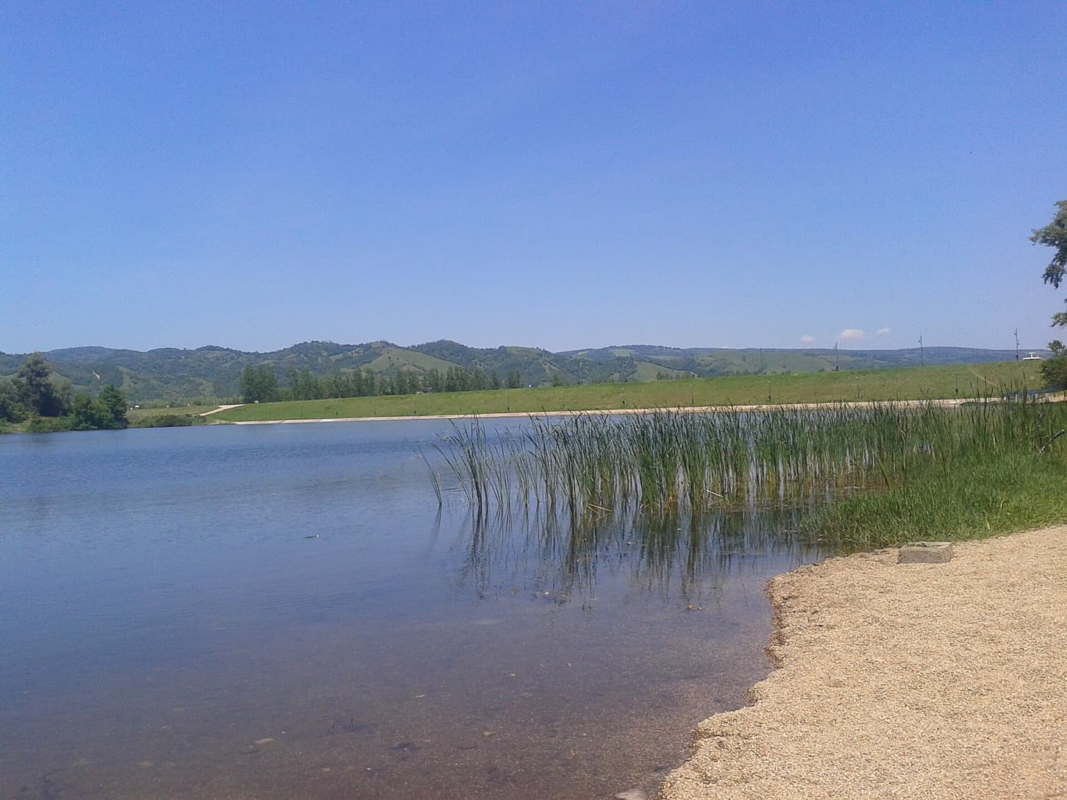 Mirna voda Srebrnog jezera sa trskom, šljunkovitom obalom i zelenim brdima
